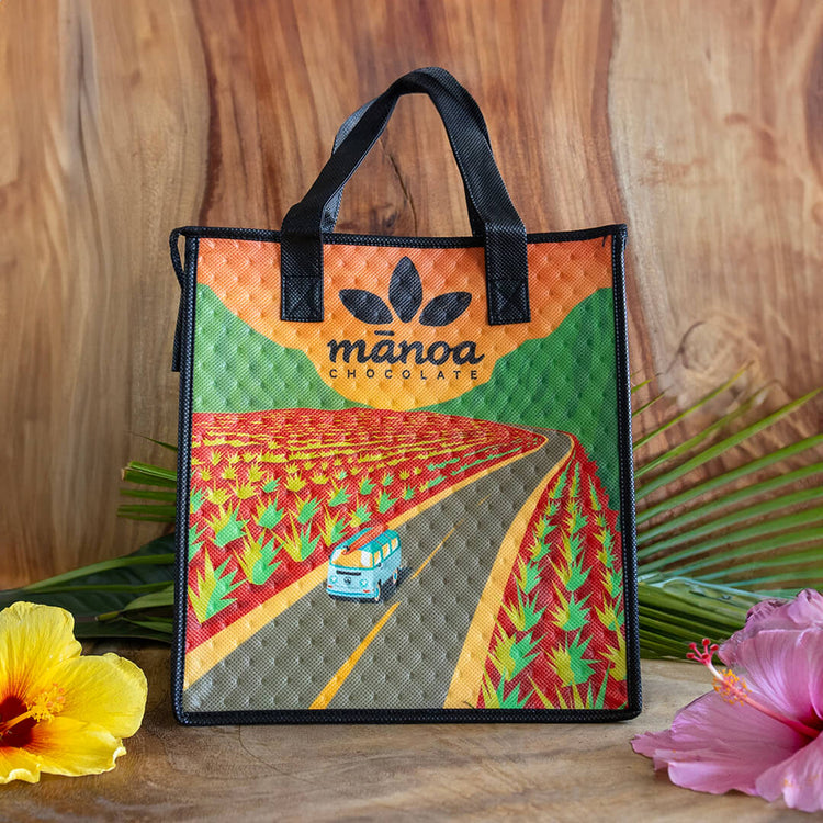 Image of a resuable Manoa Chocolate shopping bag with a graphic of a Volkswagen van driving on a tropical road on a wooden table with tropical plant garnishes
