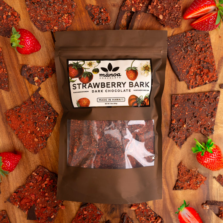 Image of Strawberry Bark Dark Chocolate package surrounded by Strawberry Bark pieces and freshly cut strawberries on table.