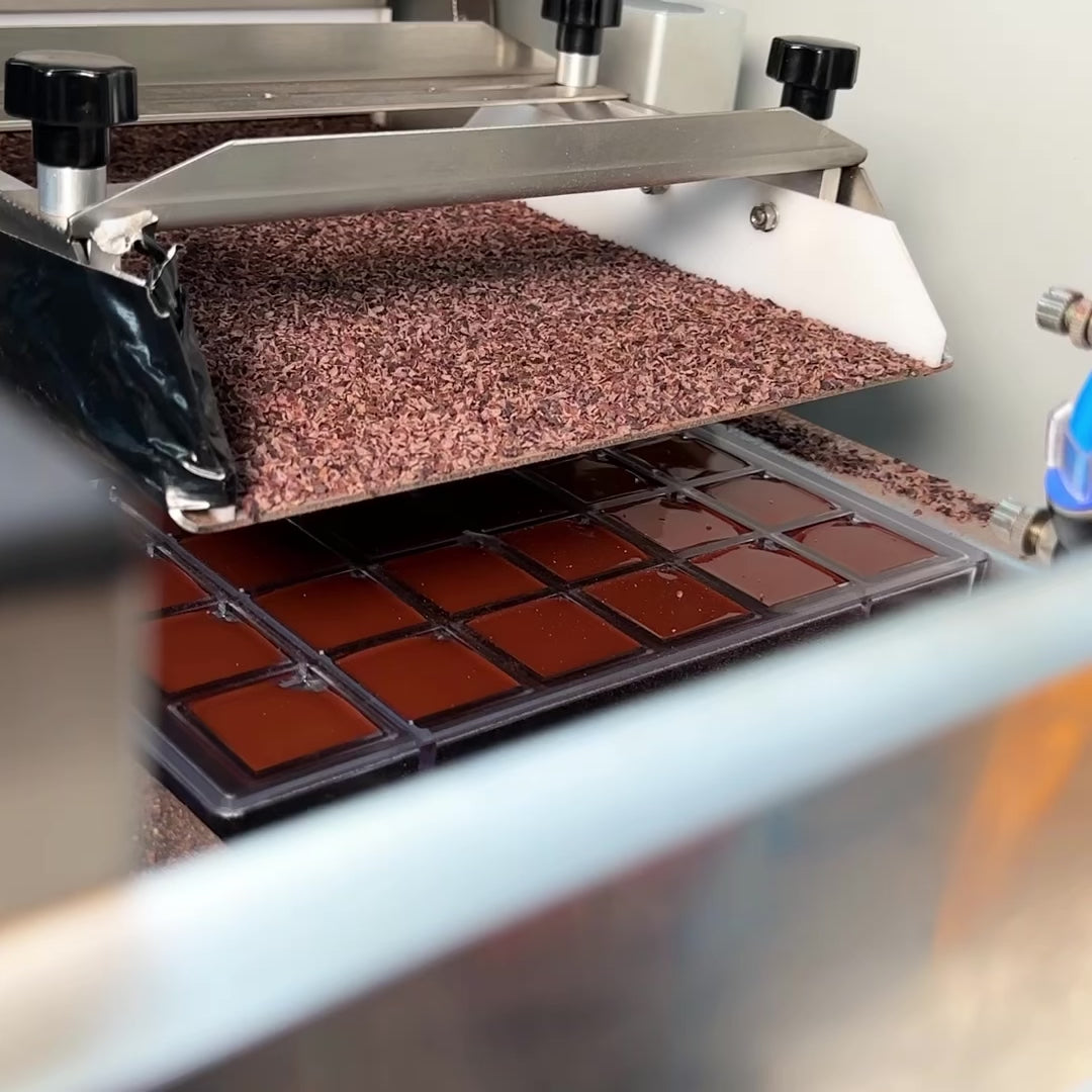 Video of product Breakfast Bites being created, with cacao nibs being poured onto the back of small chocolate squares, then those squares being removed from a mold, and finally displayed