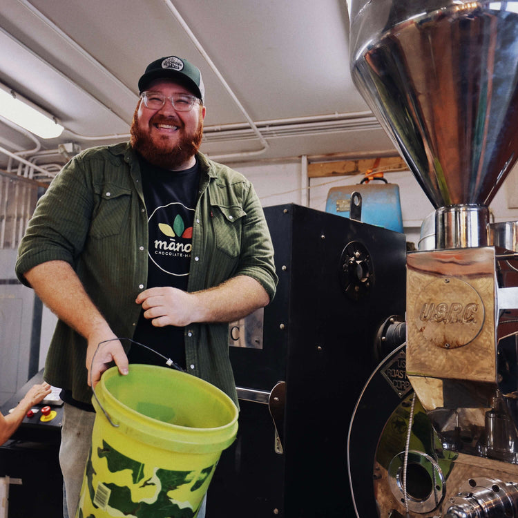Man holding a green bucket next to a coffee roasting machine in a workshop setting.