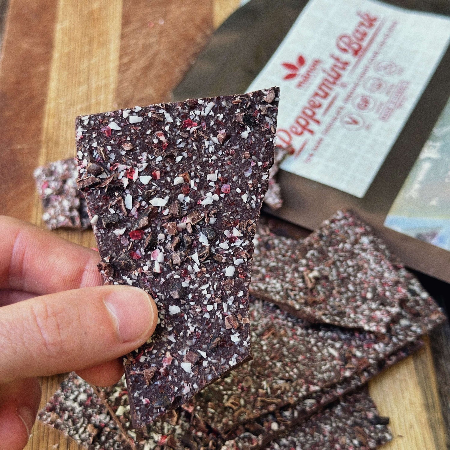 Hand holding a dark chocolate peppermint bark with a bag of peppermint bark in the background.