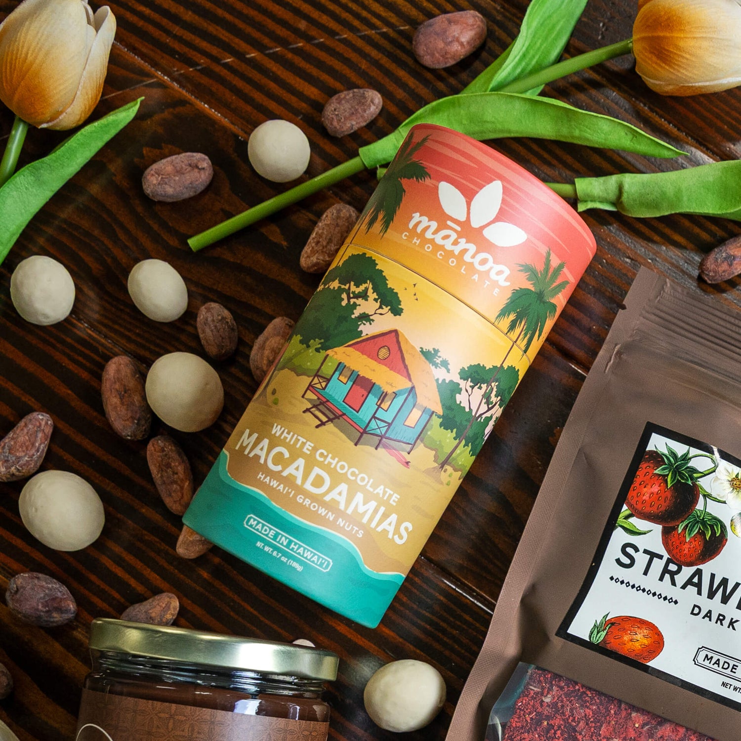 Image of a tube of White Chocolate Macadamias surrounded by cacao seeds, white chocolate macnuts, and tulips.