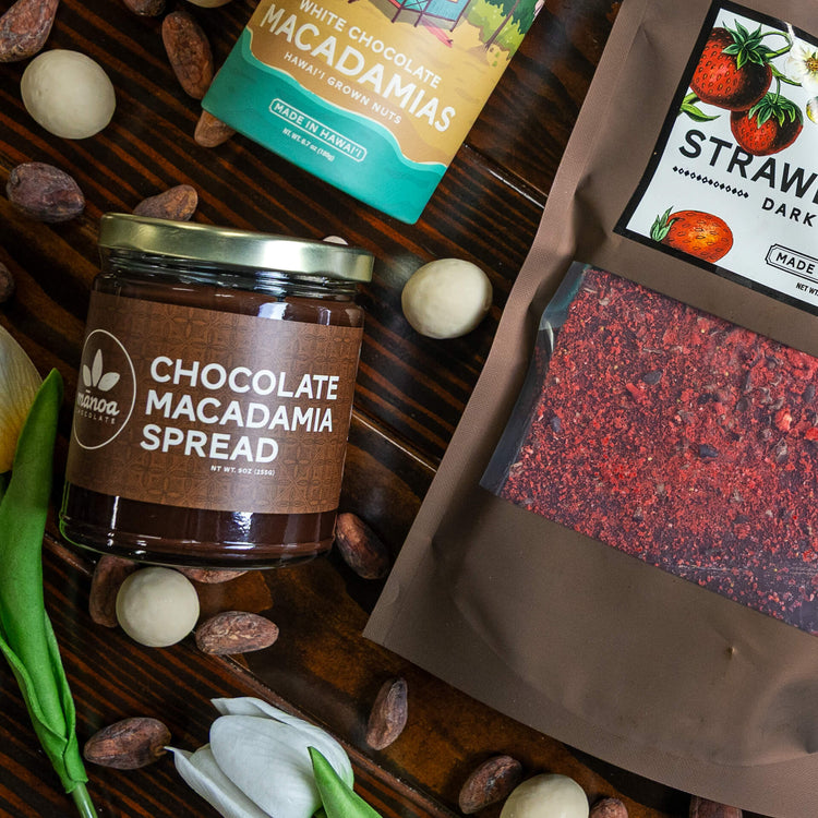 Image of Chocolate Macadamia Spread jar, a tube of White Chocolate Macadamias, and a bag of Strawberry Bark garnished with cacao seeds, white macnuts, and tulips.