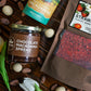 Image of Chocolate Macadamia Spread jar, a tube of White Chocolate Macadamias, and a bag of Strawberry Bark garnished with cacao seeds, white macnuts, and tulips.