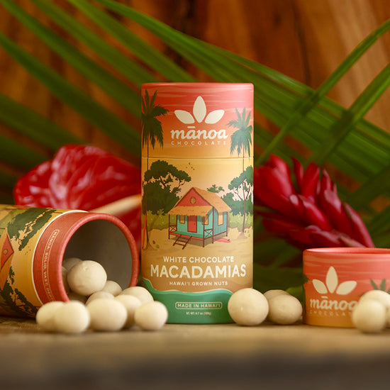 Image of white chocolate macadamia nuts scattered on table next to colorful tube package, with tropical foliage in the background