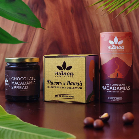 image of chocolate macadamia spread, flavors of hawaii mini box, and dark chocolate macadamia tubes on a wood table with tropical leafs in the background and foreground