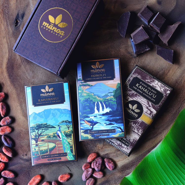kamananui, honolii, and kahaluu chocolate bar packages on a wooden table with manoa branded gift box, cacao beans, chocolate, and a banana leaf