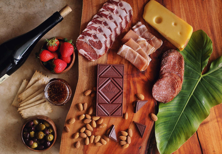Image of a chocolate bar displayed on a wooden serving board next to cured meats, strawberries, olives, jam, crackers, cheese, and a bottle of wine