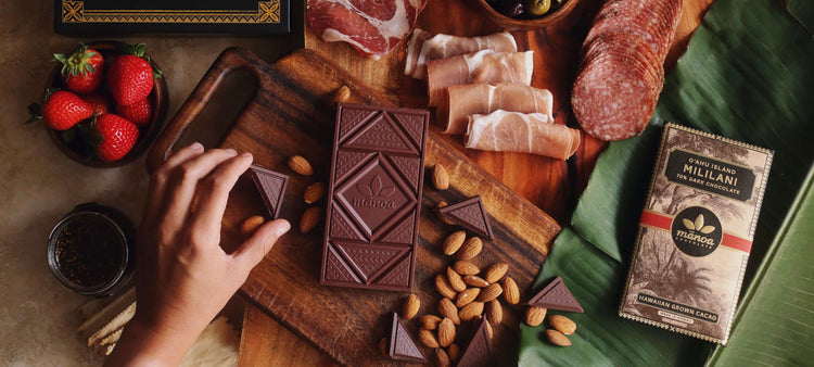 Image of a chocolate bar displayed on a wooden serving board next to cured meats, strawberries, olives, jam, crackers, cheese, and a bottle of wine