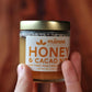 Jar of Manoa Chocolate Honey & Cacao Nibs held by a hand against a blurred wood background