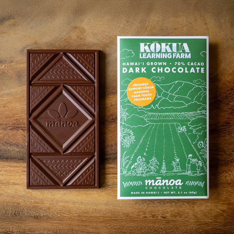 Image of a chocolate bar with a green package that says "Kokua Learning Farm Hawaii Grown 70% cacao dark chocolate" laying on a wooden surface. A sticker on the package says "proceeds support kokua learning farm youth programs"