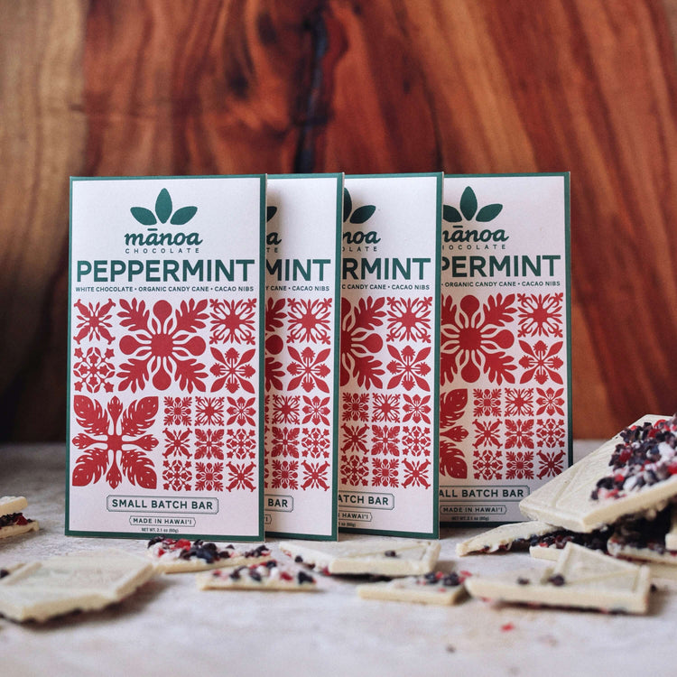 image of four packages of white Manoa Chocolate Peppermint crunch bars on a wooden surface with some bars visible.