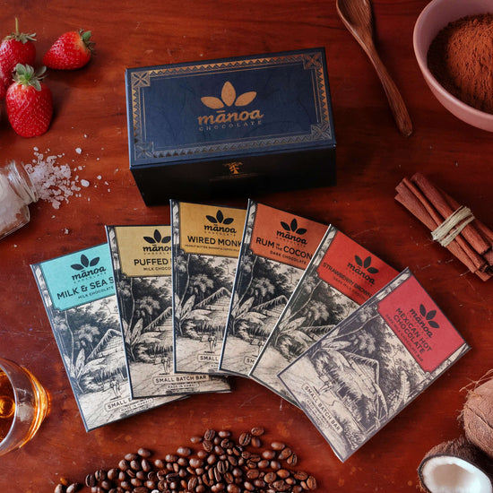 Manoa chocolate bars on a wooden surface with strawberries, coffee beans, and spices.