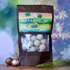 Image of a stand-up pouch of White Chocolate Macadamias, featuring a tropical beach illustration on the label and a clear window showing the white chocolate spheres inside. The pouch is displayed on a wooden surface with a whole macadamia nut and a palm leaf against a blurred tropical background.