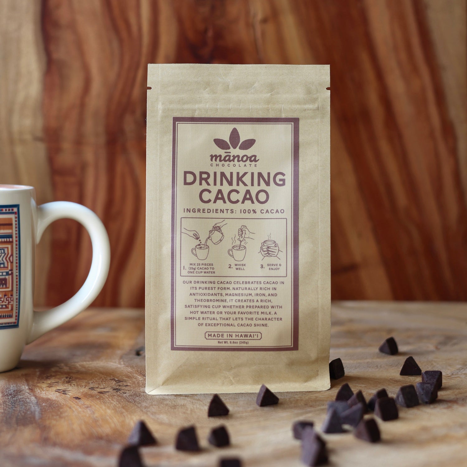 Image of a stand-up pouch of Manoa Drinking Cacao with scattered cacao pieces in front and a white mug partially visible on the left, against a warm wood plank background.
