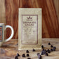 Image of a stand-up pouch of Manoa Drinking Cacao with scattered cacao pieces in front and a white mug partially visible on the left, against a warm wood plank background.