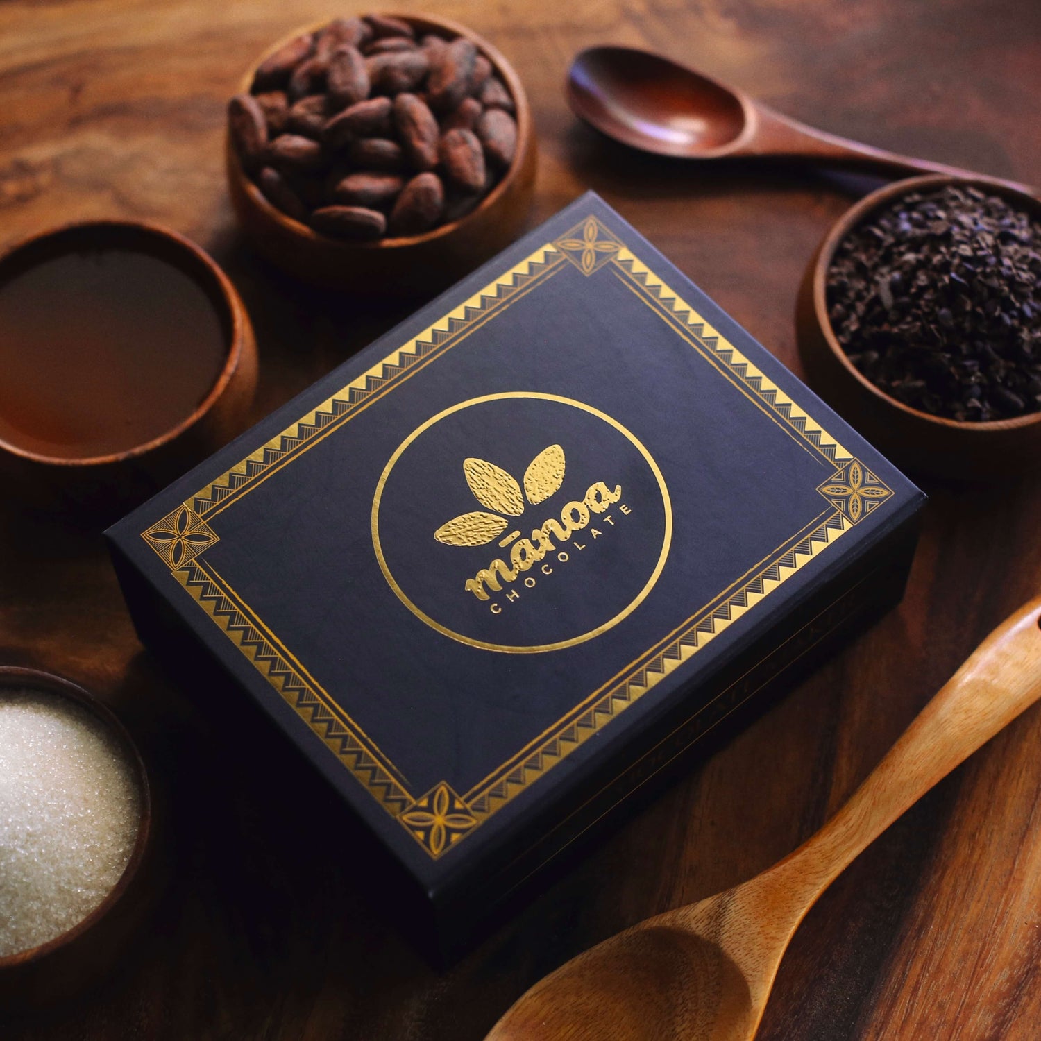 Image of gift box with gold Manoa logo sitting among cacao beans, nibs and sugar